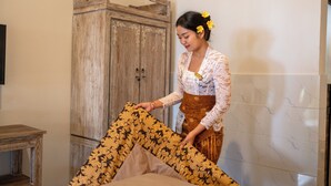Una persona colocando una manta estampada sobre una cama en una habitación con un armario de madera y paredes con azulejos.