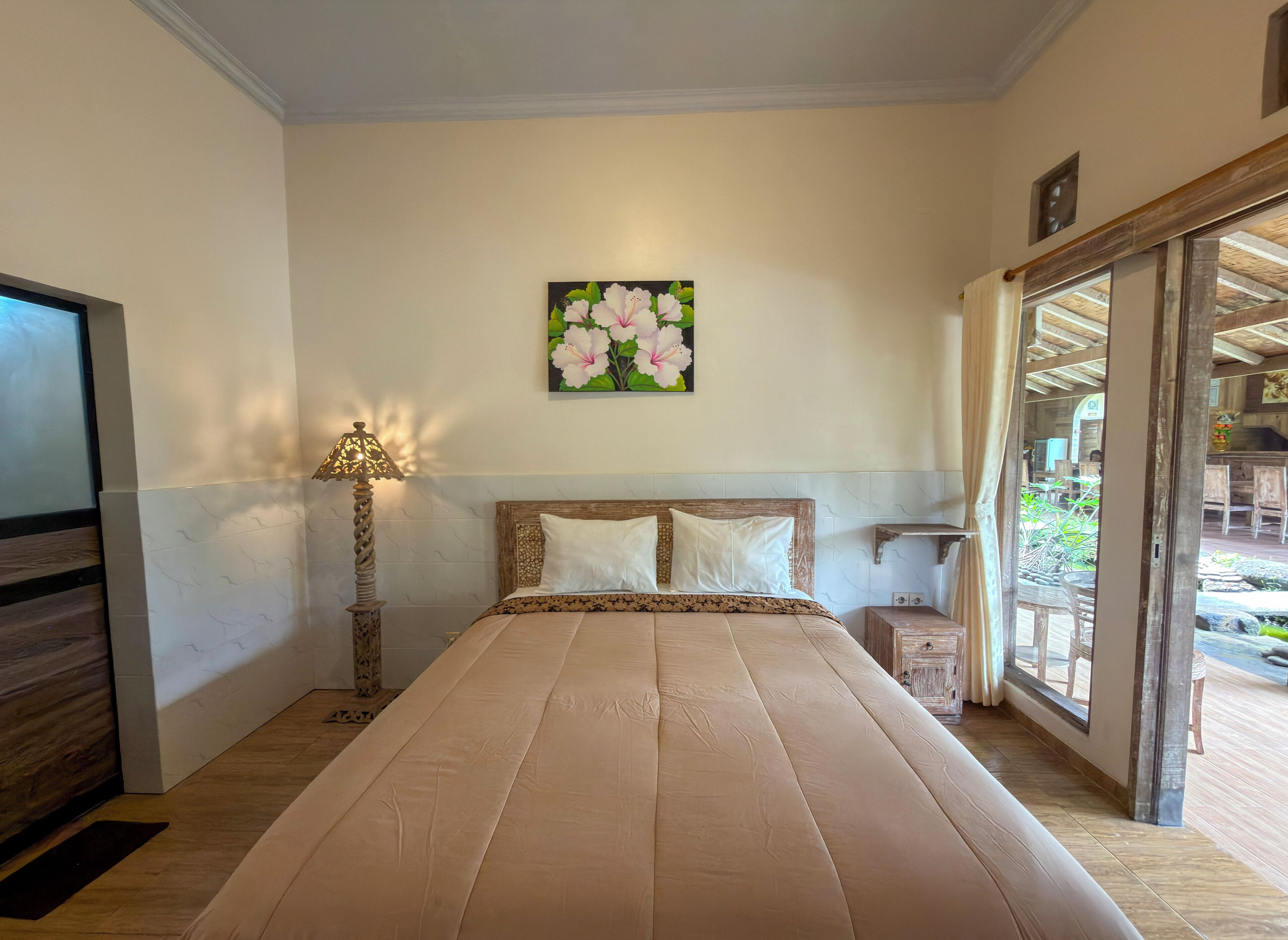 Un dormitorio con cama, mesita de noche, lámpara, cuadro y vistas al exterior.