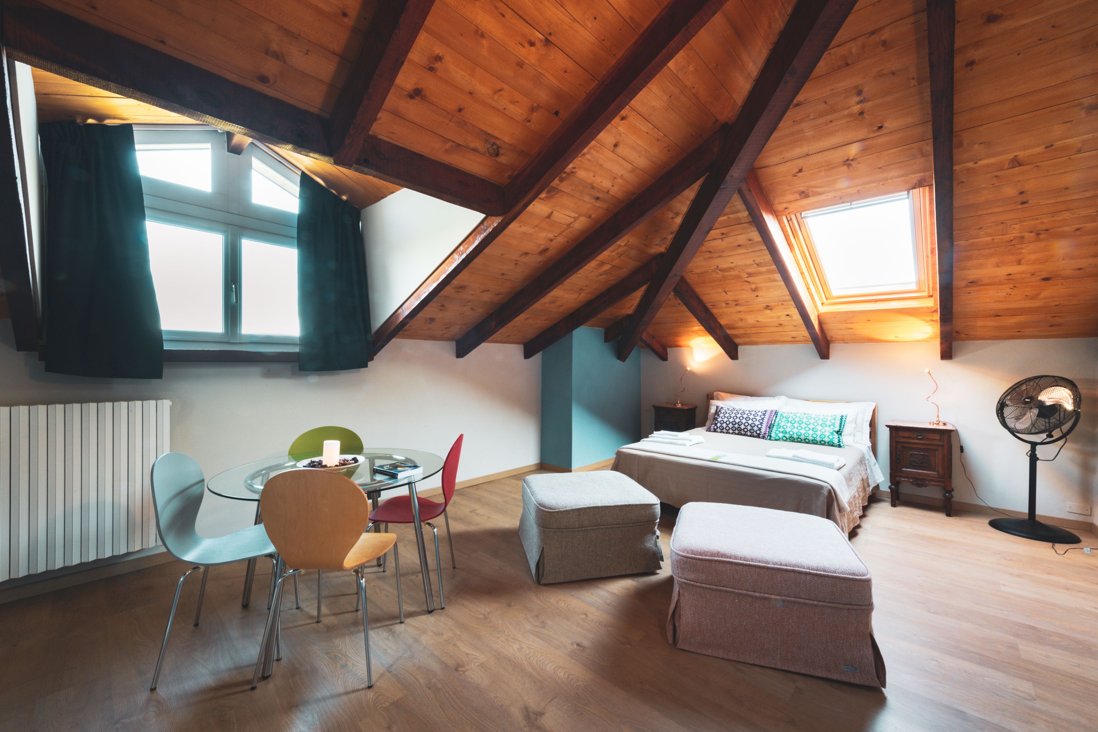 A room with a sloped ceiling, a bed, a table with chairs, and a standing fan.