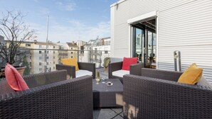 A rooftop terrace with wicker furniture, a table with a bottle and glasses, and a view of the cityscape. - Hapimag Resort München (Munich)