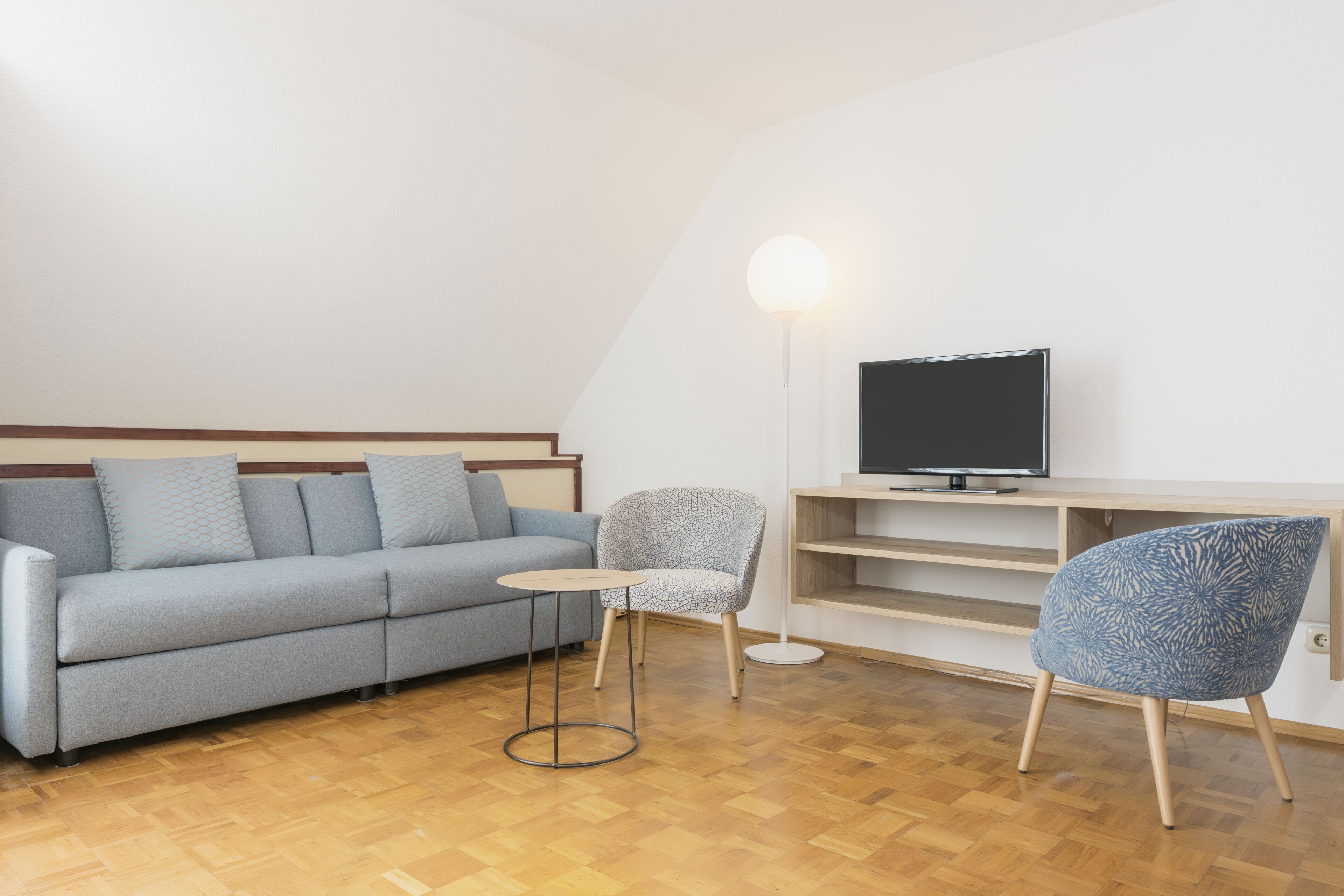 Una sala de estar moderna con un sofá, dos sillones estampados, una mesita redonda pequeña, un televisor sobre un mueble bajo y una lámpara de pie.