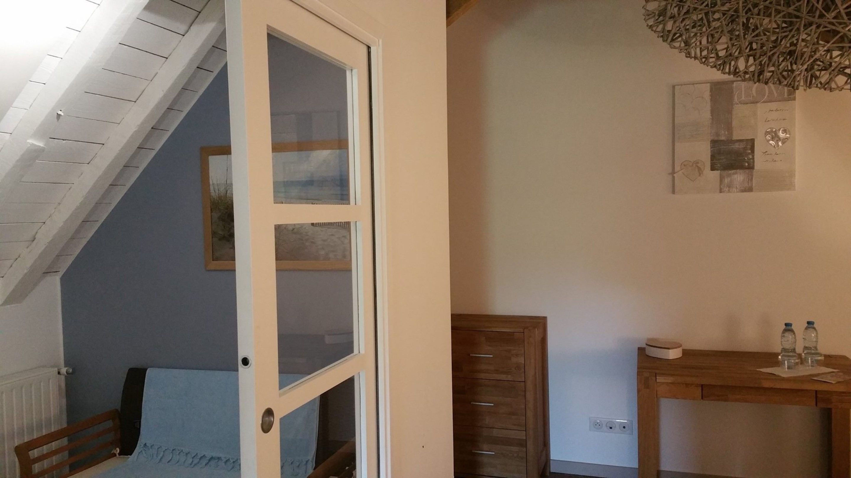 Una habitación con techo inclinado, una cama, un aparador de madera, una mesa con botellas y un cuadro en la pared.