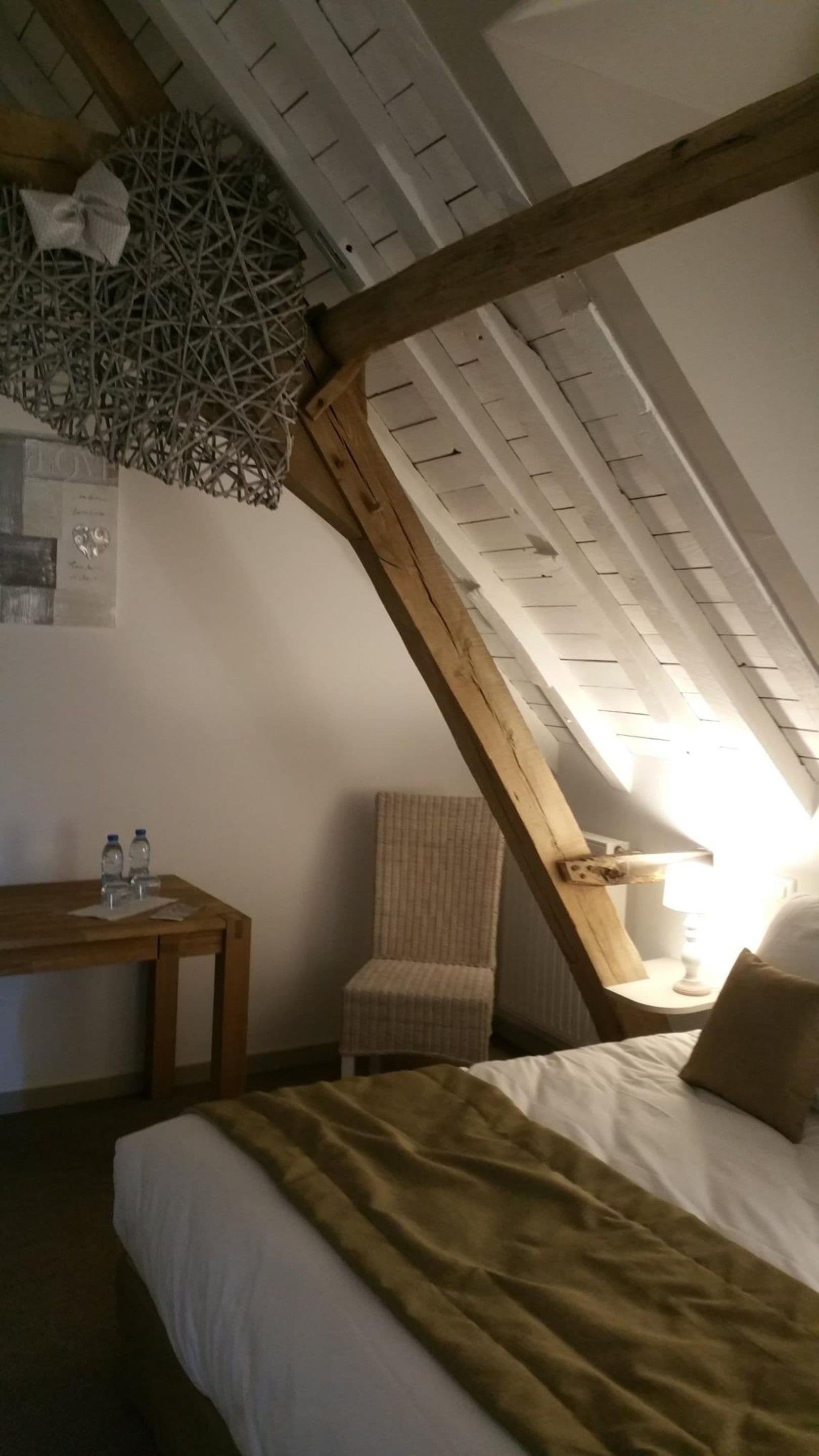 A bedroom with a sloped ceiling, a bed with a brown blanket, a chair, a small table with water bottles, and a lamp.