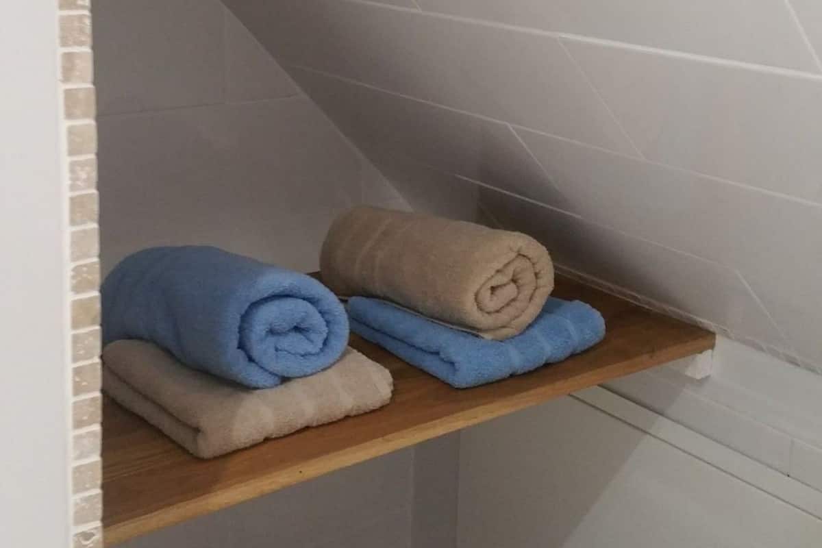 A bathroom with a wooden shelf holding rolled towels in blue and beige.