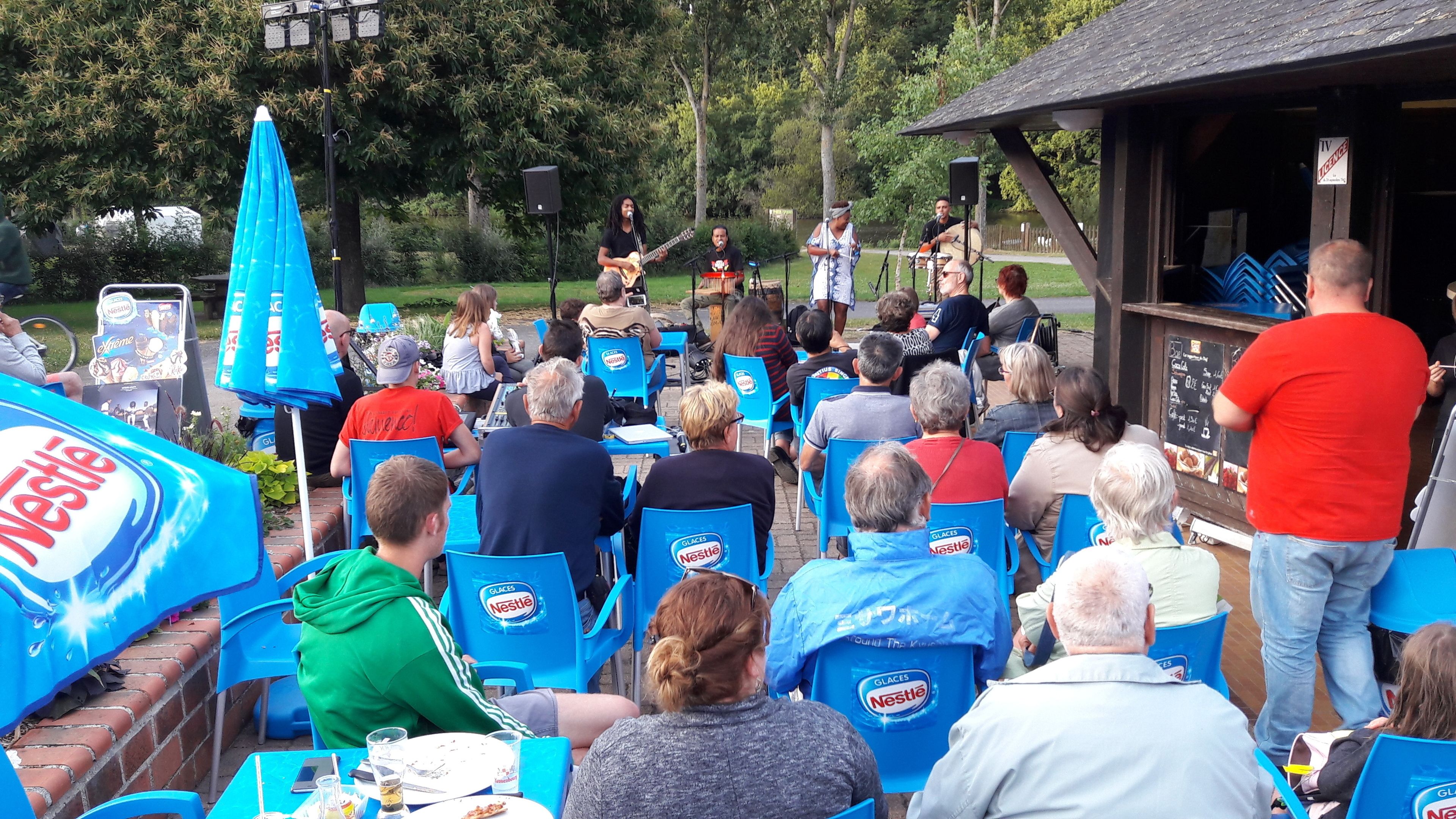An outdoor event with a stage and audience seated at tables with blue chairs.