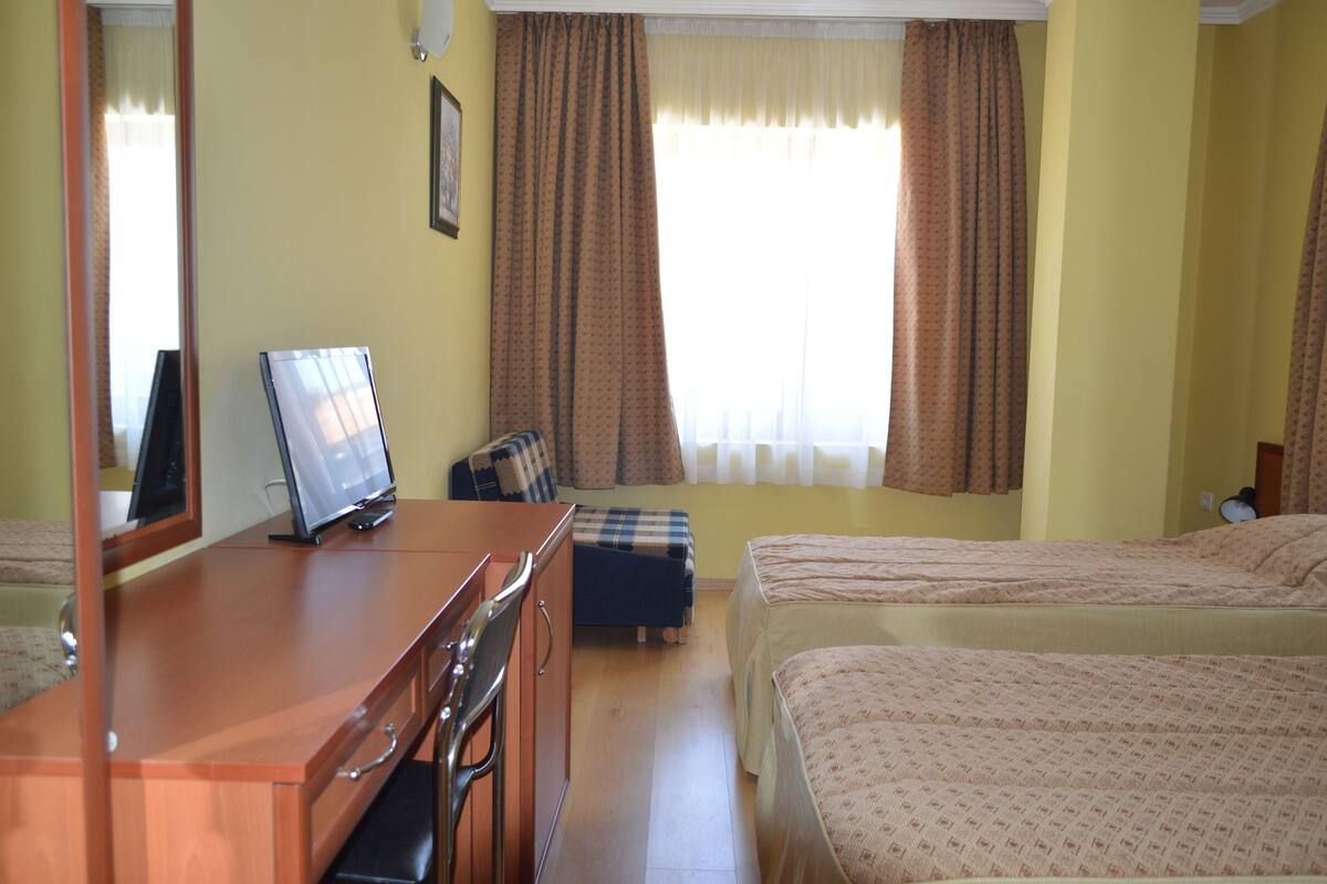 A hotel room with two beds, a wooden desk with a computer, a chair, and a window with curtains.