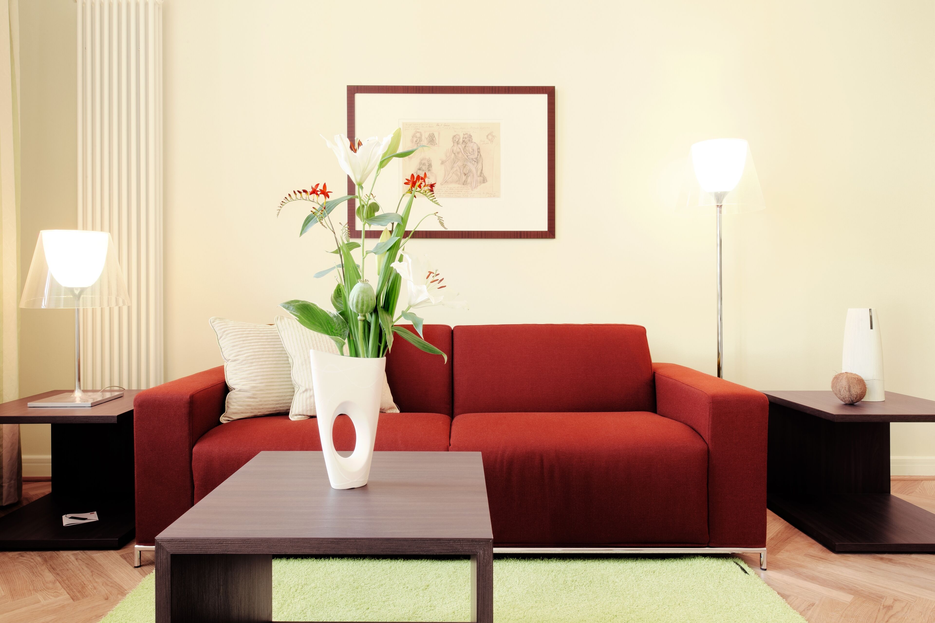 A modern living room with a red sofa, a coffee table, a white vase with flowers, and two side tables with lamps.