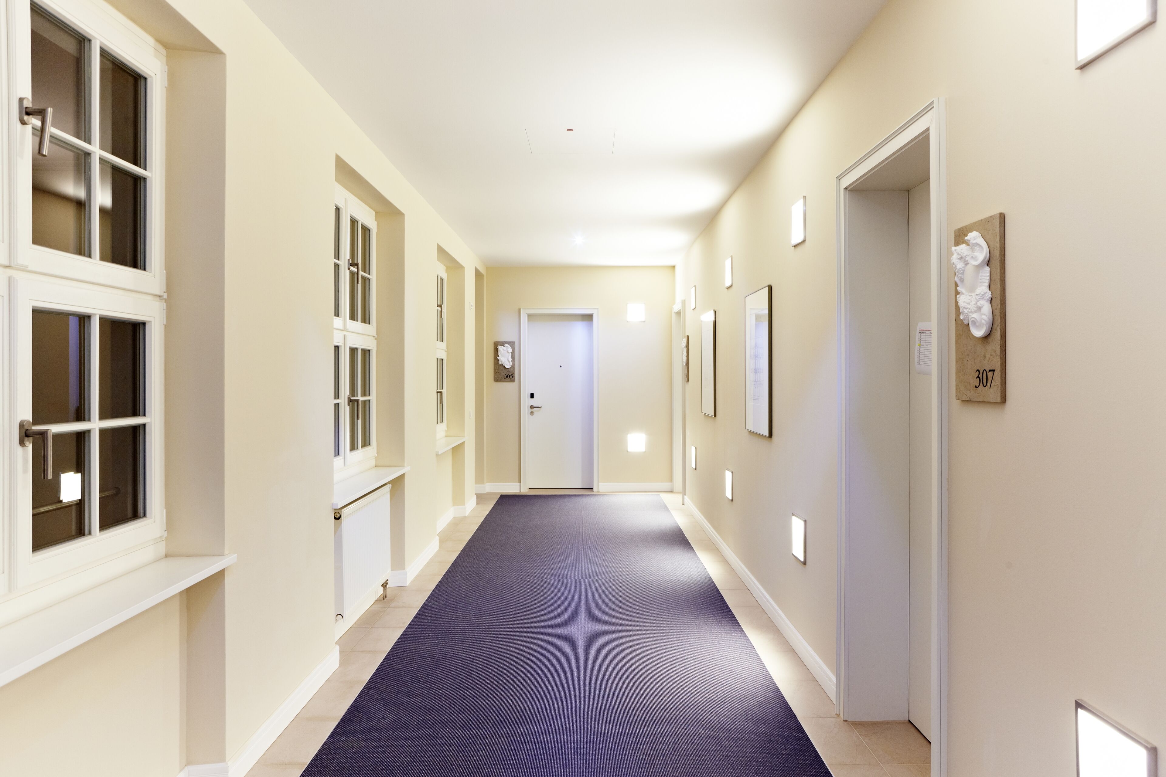Un couloir d’hôtel avec un tapis bleu, des murs de couleur beige et des portes numérotées.