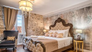 A bedroom with a large bed, a chair, a chandelier, and a stone wall. - Plaza Marchi Old Town - MAG Quaint & Elegant Boutique Hotels (Split)