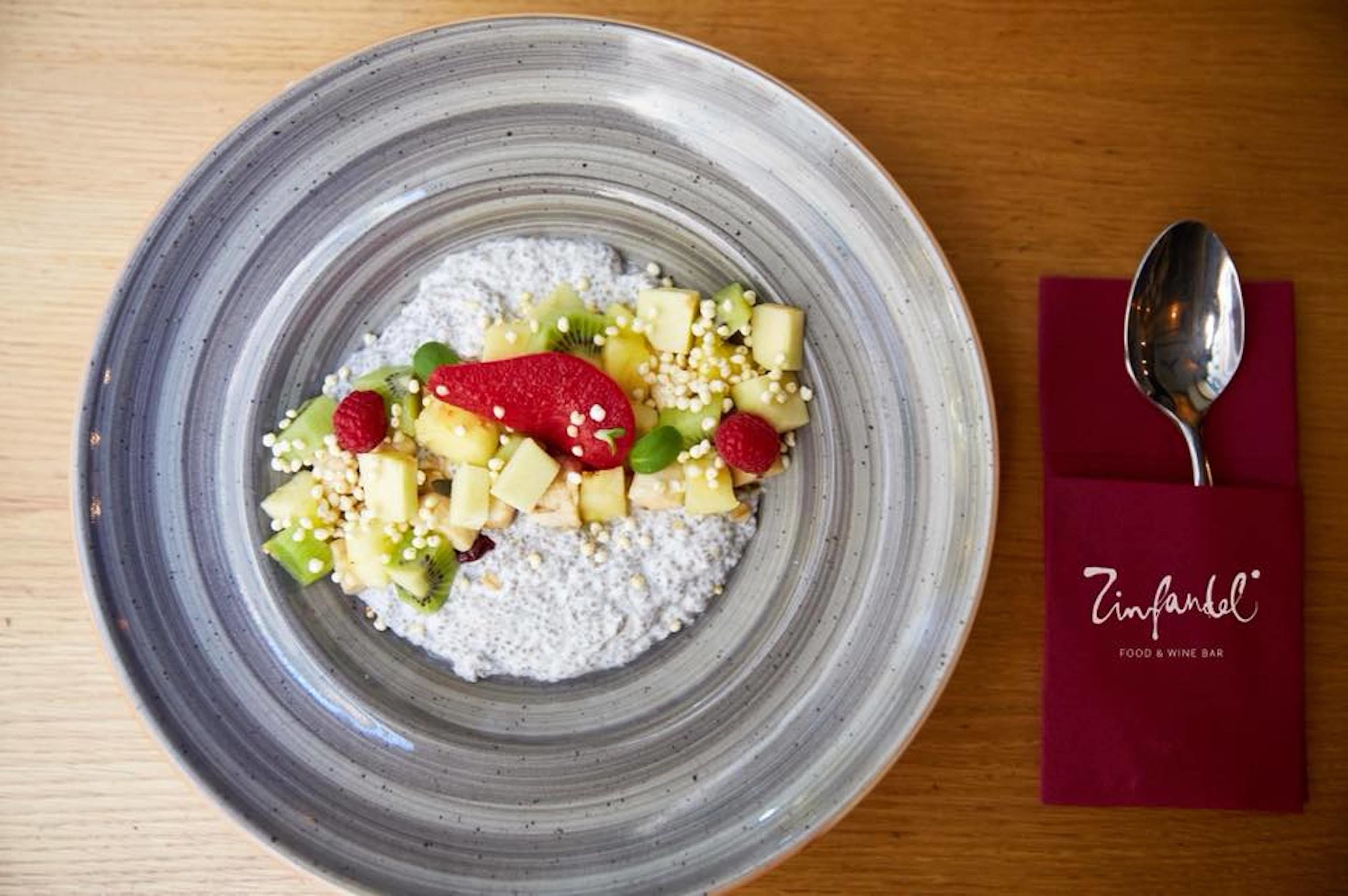 A bowl of oatmeal topped with fruits and seeds, a spoon, and a napkin with a logo on a wooden table.