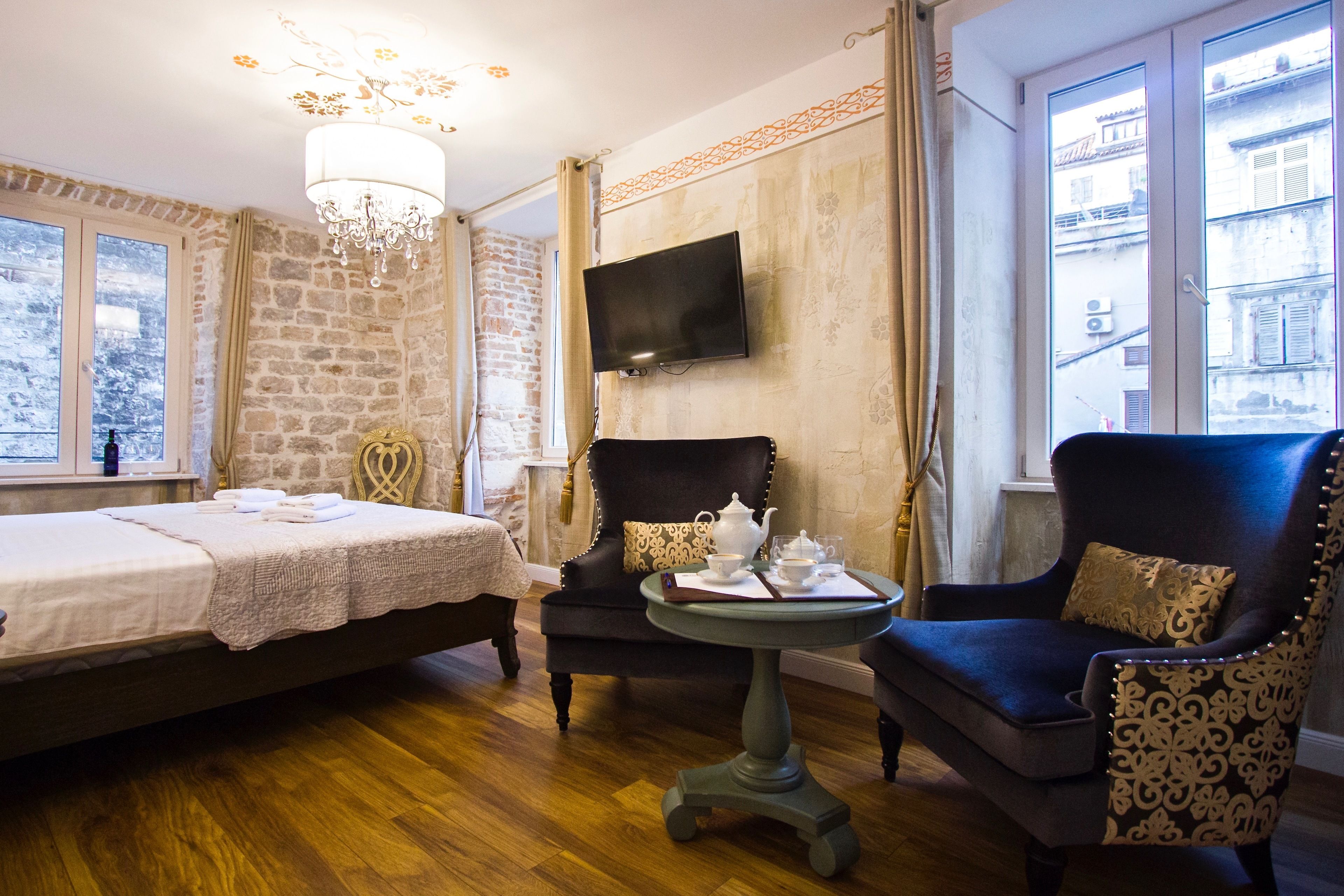 Une pièce comprenant un lit, deux fauteuils, une petite table avec une théière et des tasses, un lustre, un téléviseur à écran plat fixé au mur et une fenêtre donnant sur des immeubles.