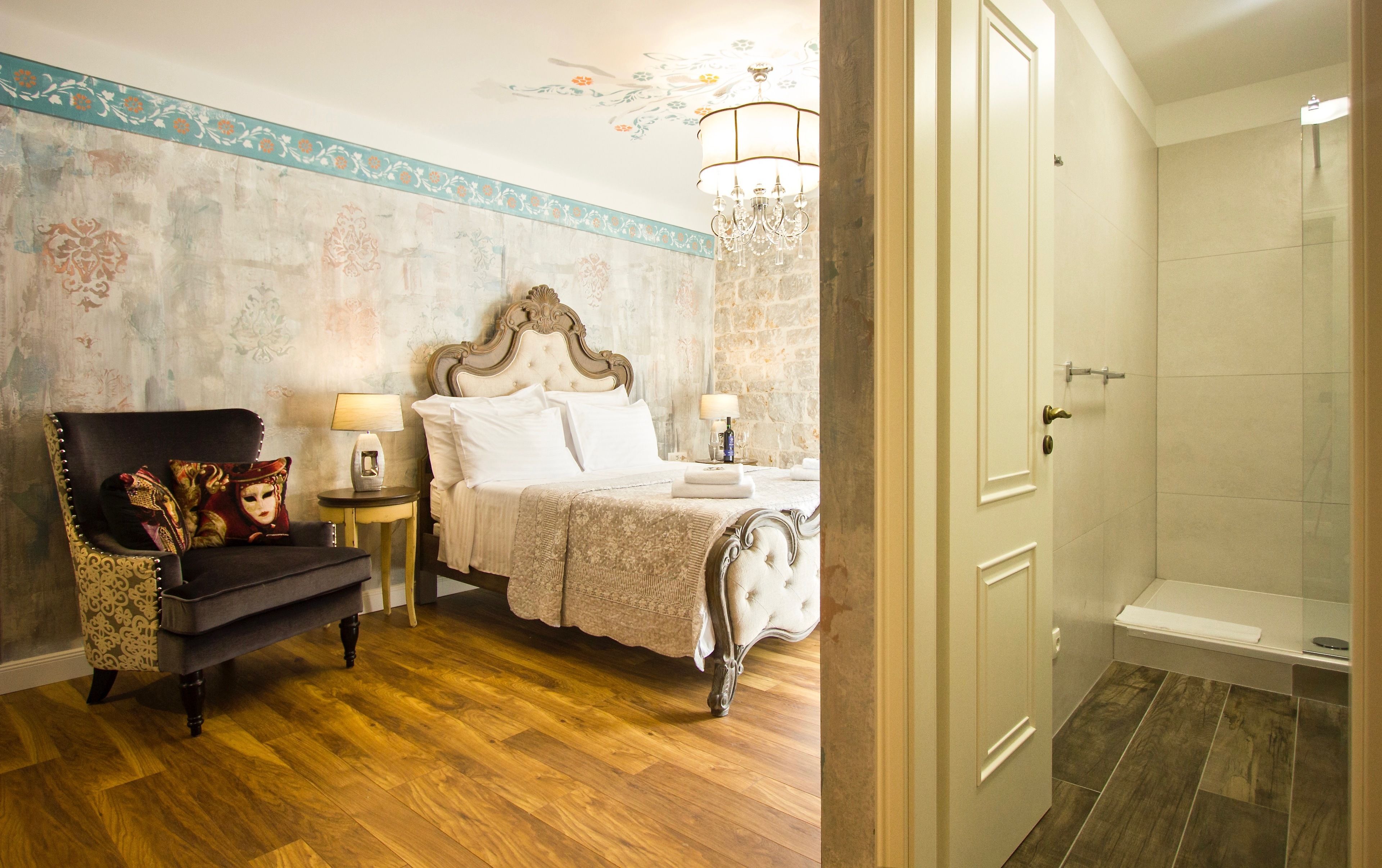 A bedroom with a bed, a chair, two bedside tables with lamps, a chandelier, and a bathroom with a shower and toilet.