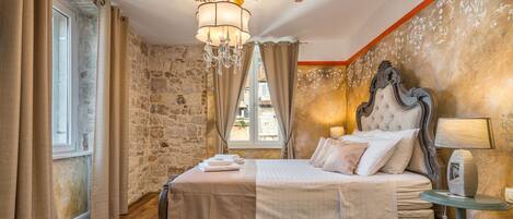 A bedroom with a stone wall, a wooden bed, a chandelier, and a window with curtains.