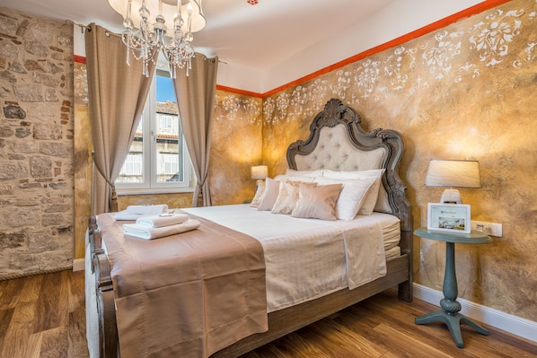 A bedroom with a large bed, a chandelier, a small table with a lamp, and a stone wall.