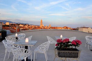 A rooftop terrace with white metal chairs and tables, a potted plant with pink flowers, and a view of a cityscape with a prominent tower.