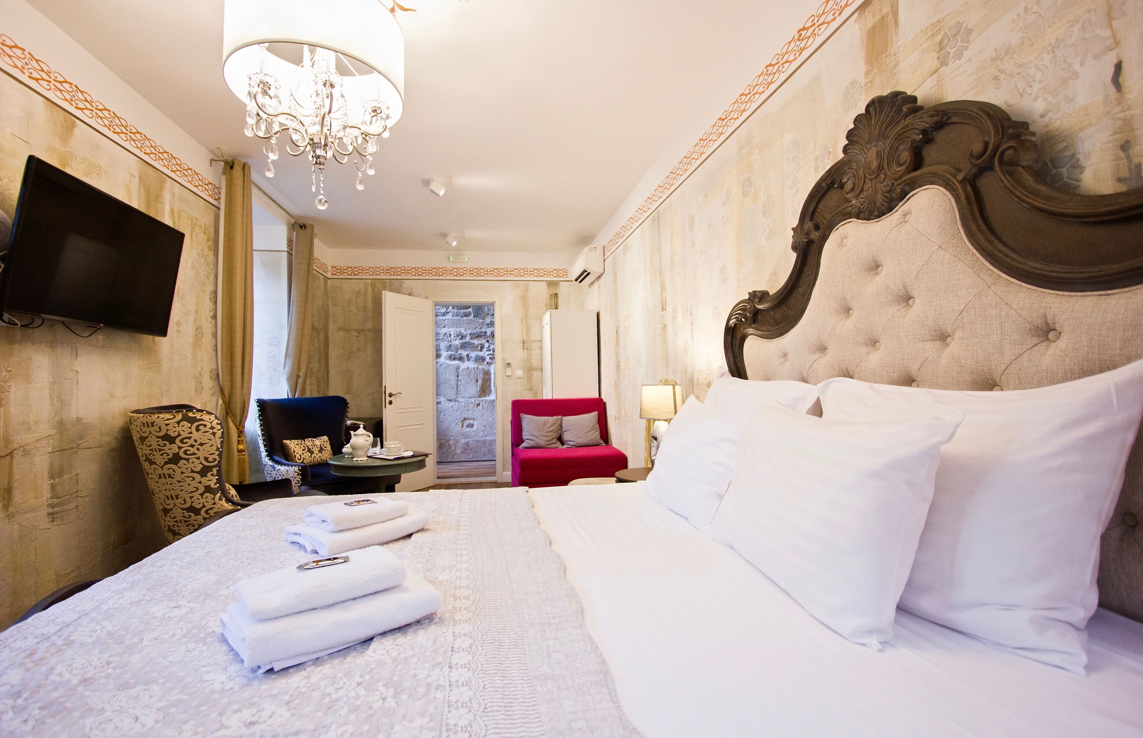 A hotel room with a large bed, a television on the wall, a chandelier, and a red chair.