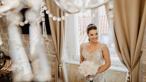A bride in a white dress with lace details, holding a bouquet, standing in a room with a large window, curtains, and a chandelier. - Plaza Marchi Old Town - MAG Quaint & Elegant Boutique Hotels (Split)
