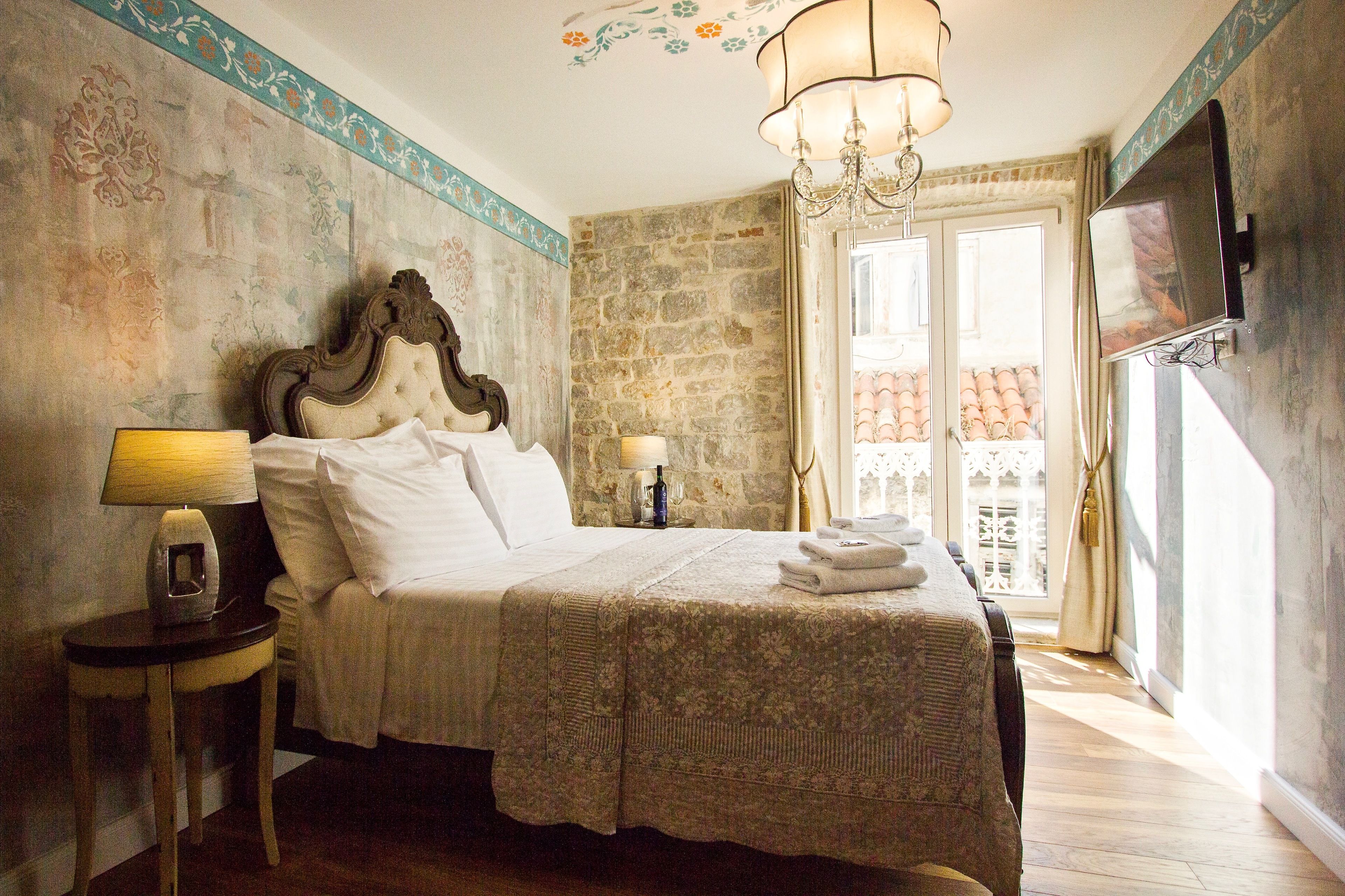A bedroom with a large bed, a nightstand with a lamp, a chandelier, and a view of a balcony with red tiles.