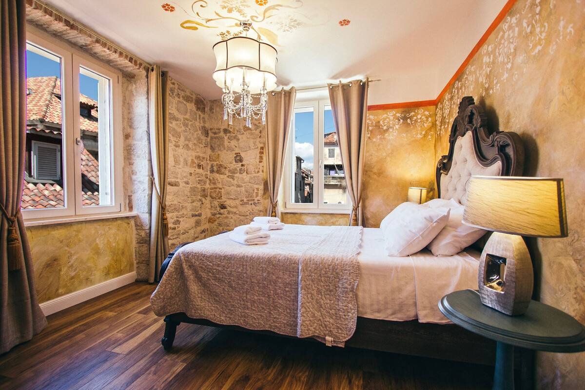 Ein Schlafzimmer mit Stehwänden, einem Bett mit Kopfteil, einer Chandelüstere, einem Fenster mit Vorhängen und einem Nachttisch mit Lampe.