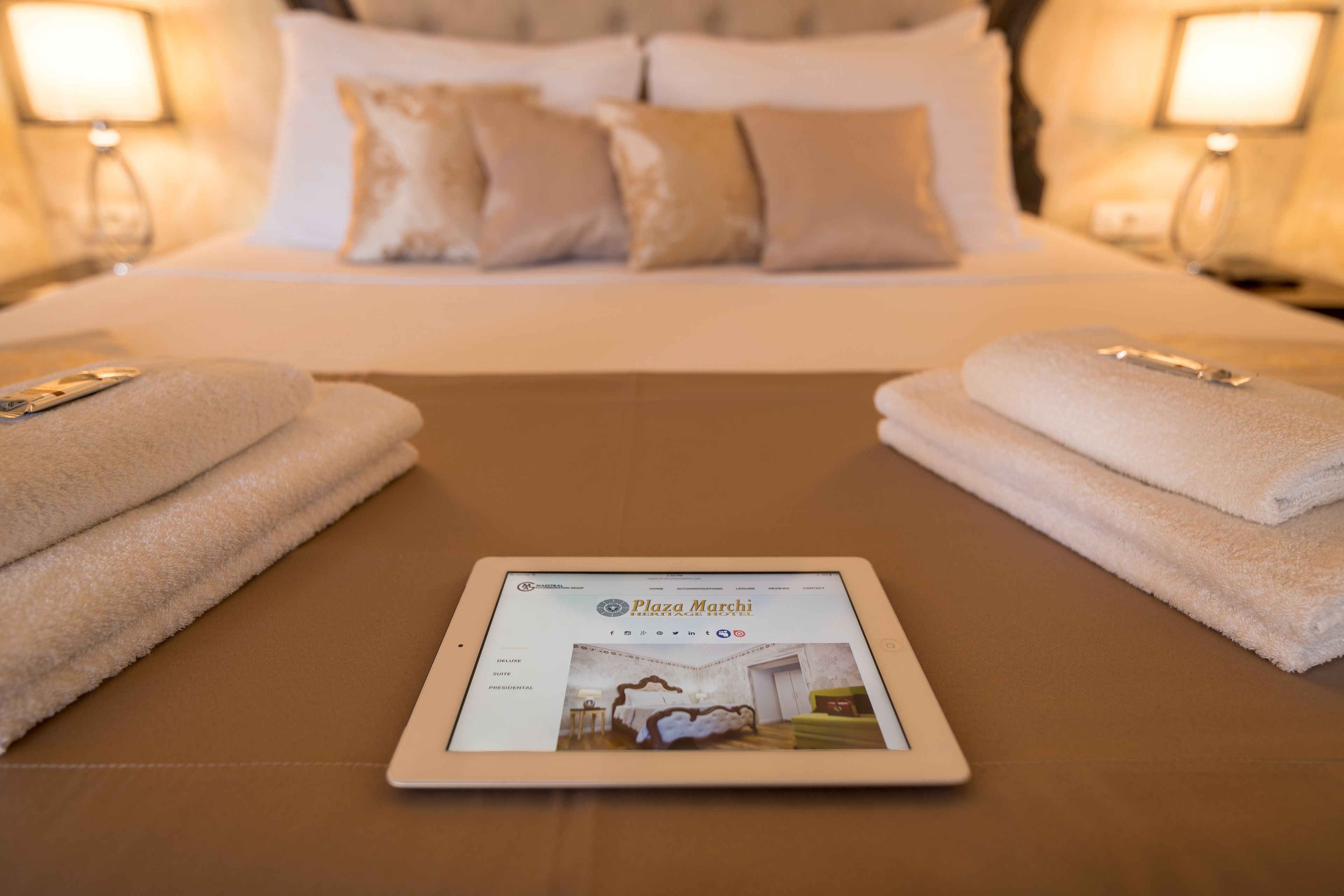 A tablet displaying a hotel website on a bed with towels and a nightstand with a lamp.