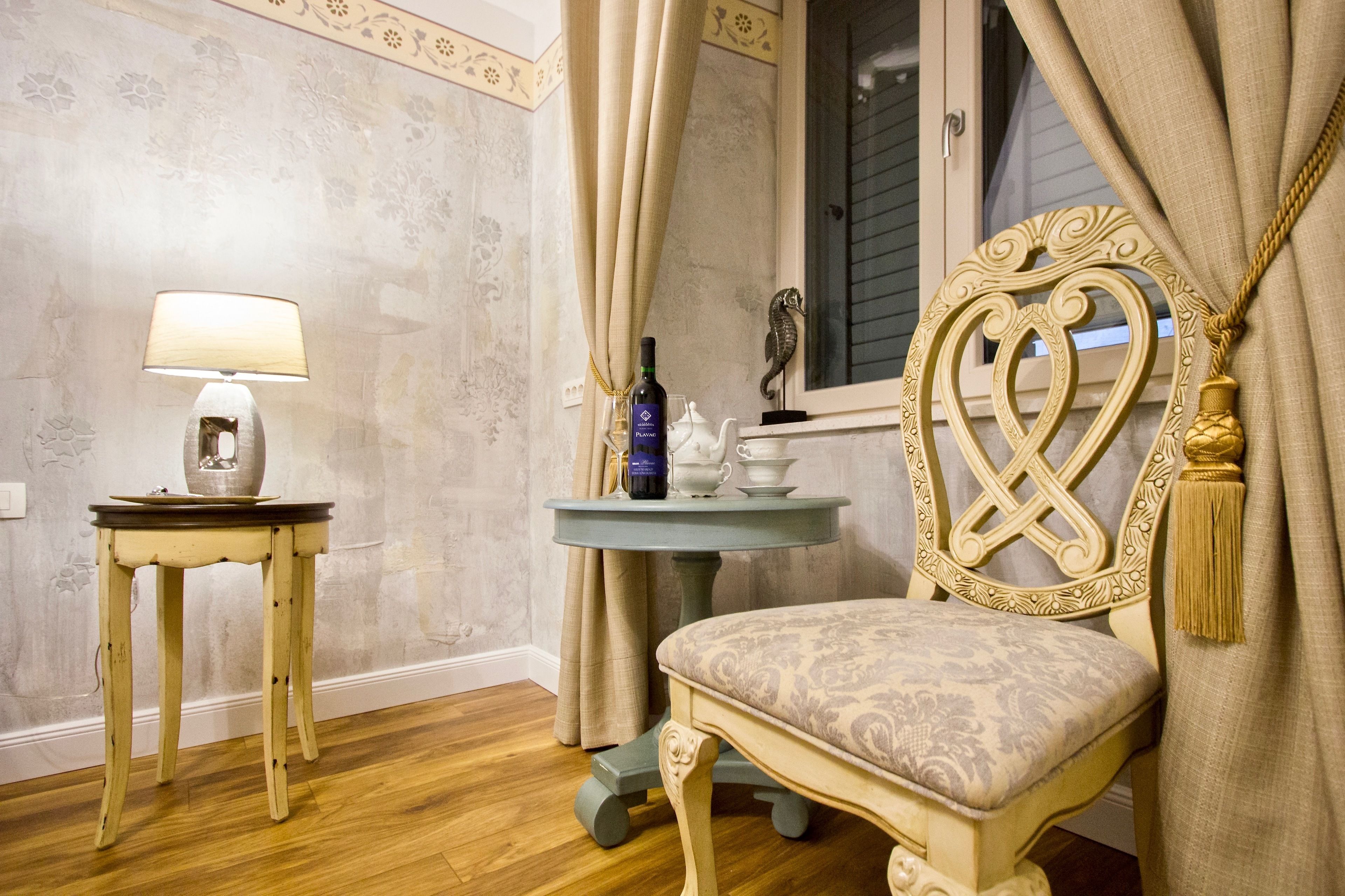 A room with a wooden floor, a table with a lamp, a chair with a heart-shaped frame, and a window with curtains.