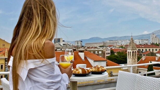 A person with long blonde hair sitting at a table with croissants and orange juice, overlooking a cityscape with buildings and a church tower. - Plaza Marchi Old Town - MAG Quaint & Elegant Boutique Hotels (Split)