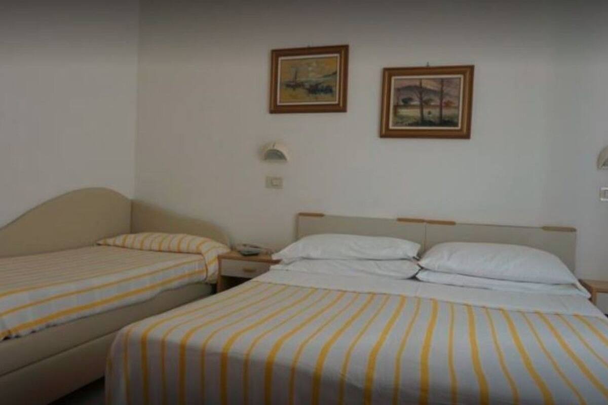 A hotel room with two single beds, each with striped bedding. There are two framed pictures on the wall, a nightstand with a lamp, and a small table.