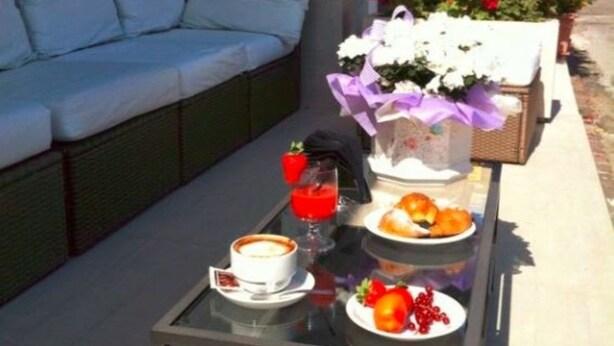 A glass table with breakfast items, a white sofa, and a flower arrangement on a balcony. - Hotel Aurora (Gabicce Mare)