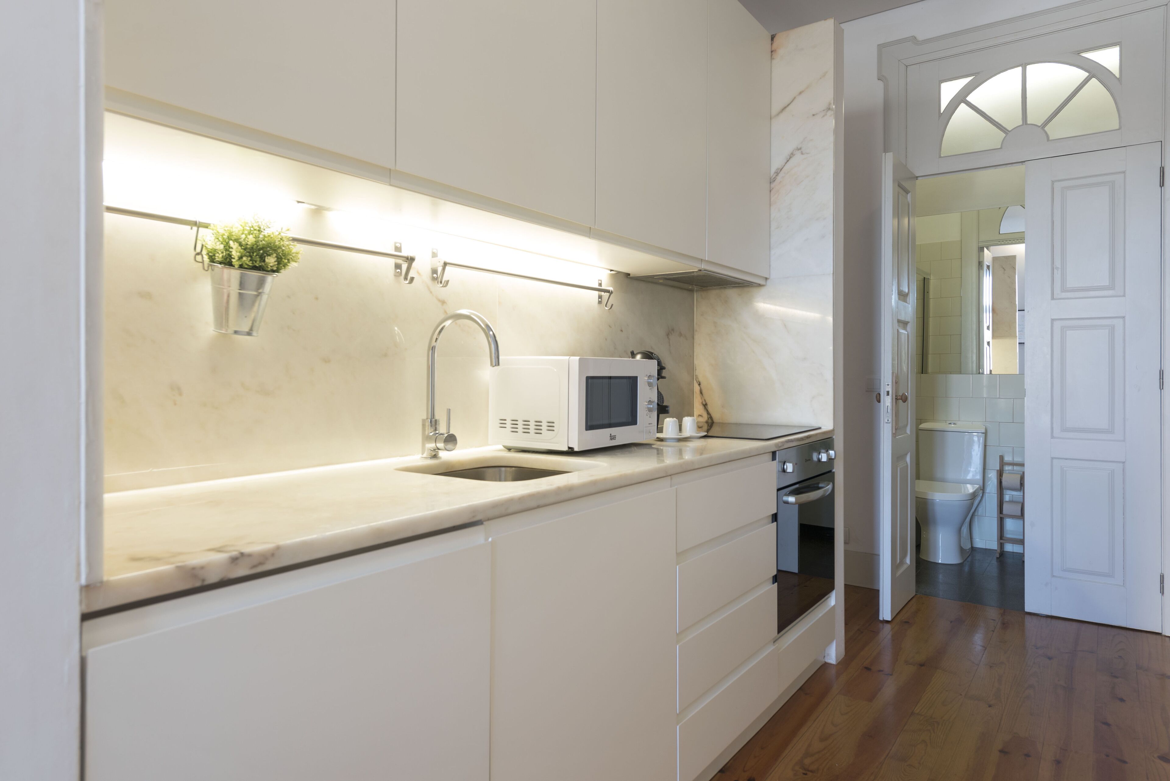 Une cuisine moderne avec des armoires blanches, un évier en acier inoxydable, un four à micro-ondes et un four traditionnel. On aperçoit une petite plante sur le plan de travail et une chaise en bois à l’arrière-plan.