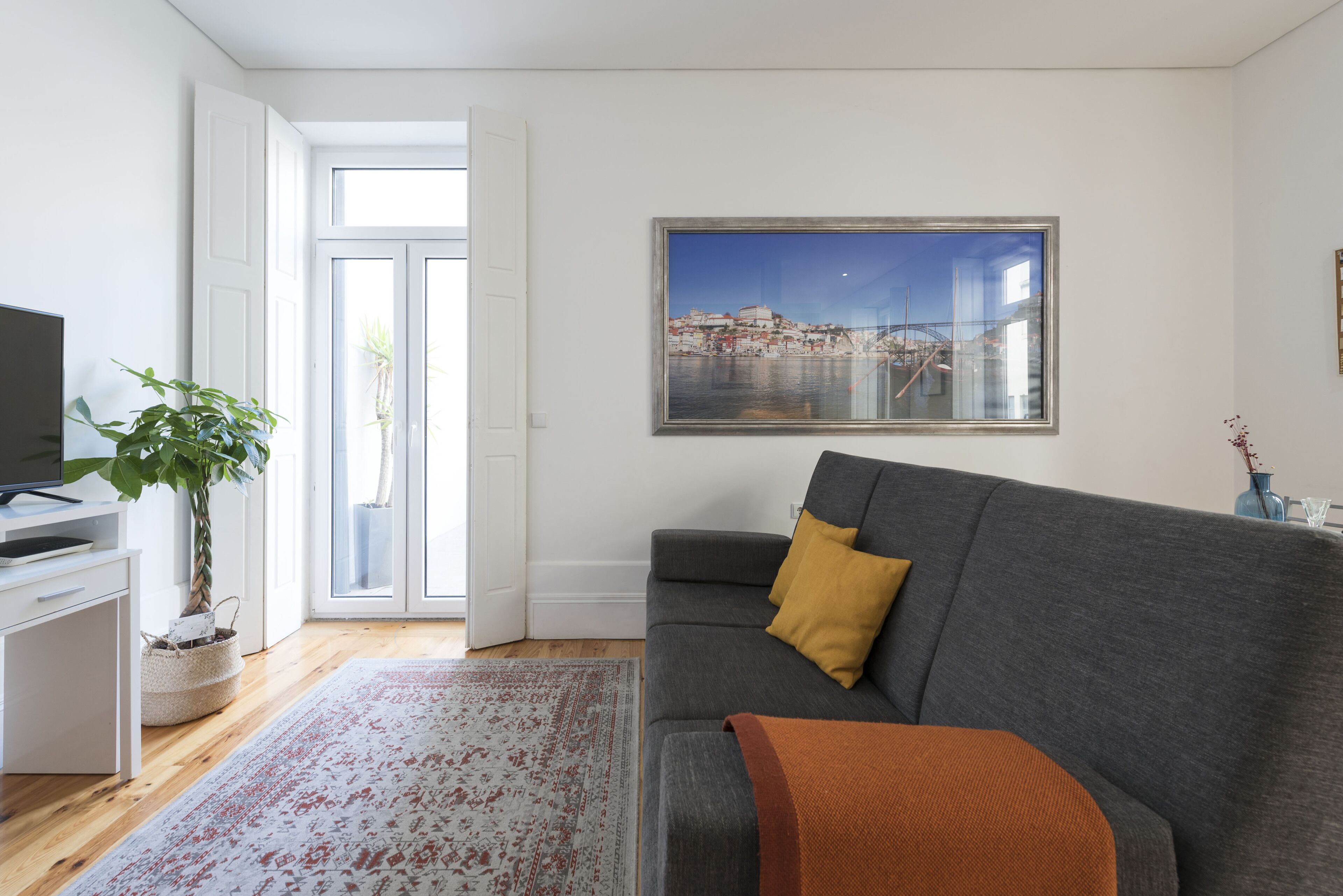 Un salon avec un canapé gris, un coussin jaune, une grande fenêtre donnant sur le paysage urbain, un téléviseur à écran plat et une plante en pot.