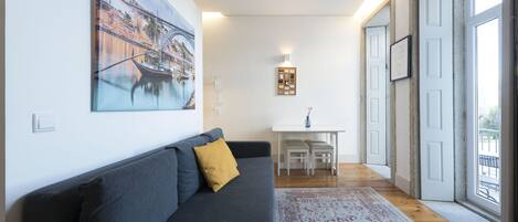 A modern living room with a sofa, a painting, a small table, and a view through large windows.