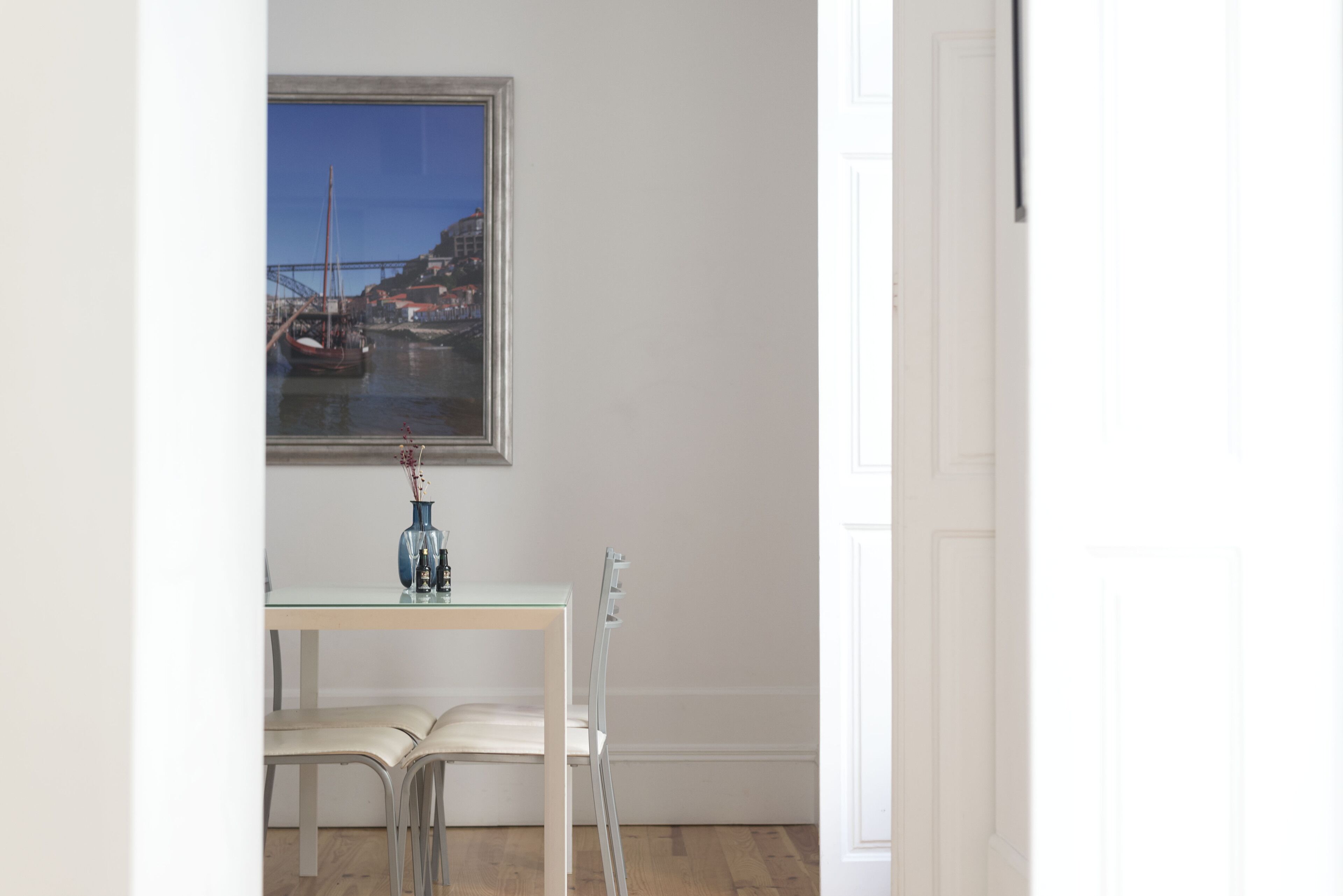 Ein Zimmer mit einer weißen Tür, einem gerahmten Gemälde eines Hafens, einem kleinen Tisch mit einer Vase und zwei weißen Stühlen.