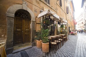 A cobblestone street with outdoor seating at a café, featuring terracotta pots with plants, wooden tables, and a stone archway entrance. - POP Art B&B (Rome)