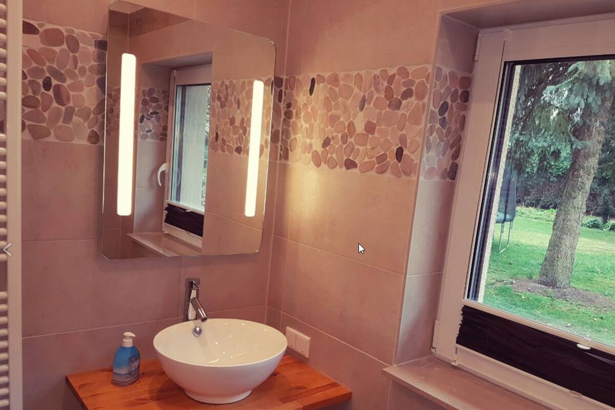 Une salle de bain avec un mur décoré d’une mosaïque de pierre, un lavabo blanc et une fenêtre donnant sur les arbres.