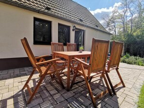 Un ensemble de table et de chaises en bois est installé sur une terrasse dallée devant une maison dont le toit est recouvert de tuiles.