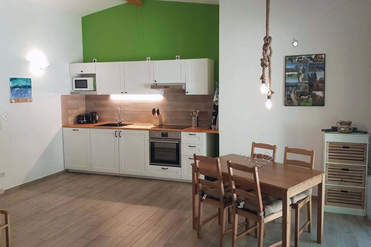 Une cuisine avec des meubles blancs, une table à manger en bois et des chaises. On y trouve un luminaire suspendu et un four à micro-ondes posé sur le comptoir.