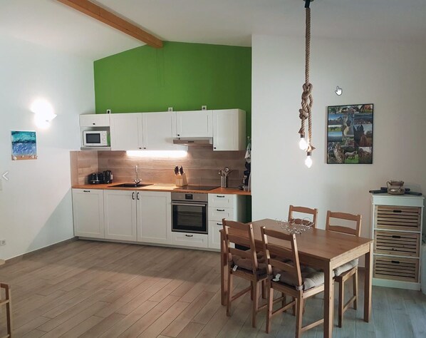Une cuisine avec des meubles blancs, une table à manger en bois et des chaises. On y trouve un luminaire suspendu et un four à micro-ondes posé sur le comptoir.