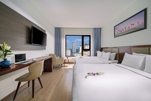 A hotel room with a large bed, a desk with a laptop, a TV mounted on the wall, and a city view through the window. - Aria Grand Hotel & Spa (Da Nang)