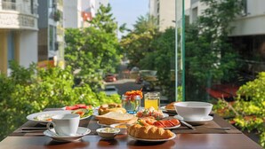 A breakfast table with plates of food, a cup, and a glass of juice, set against a backdrop of greenery and buildings.