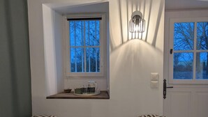 Une pièce comprenant deux fauteuils à motifs, une petite table, une fenêtre donnant sur des arbres et une porte surmontée d’un luminaire.