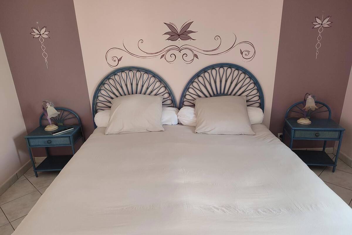 Un lit double avec une tête de lit ornée d’un motif décoratif, deux tables de chevet avec des lampes et un mur décoré de motifs floraux.