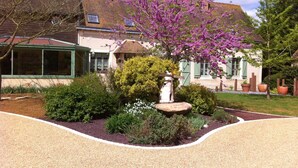 Une maison dotée d’un jardin agrémenté d’une fontaine en pierre, d’arbustes en fleurs et d’une allée gravillonnée.