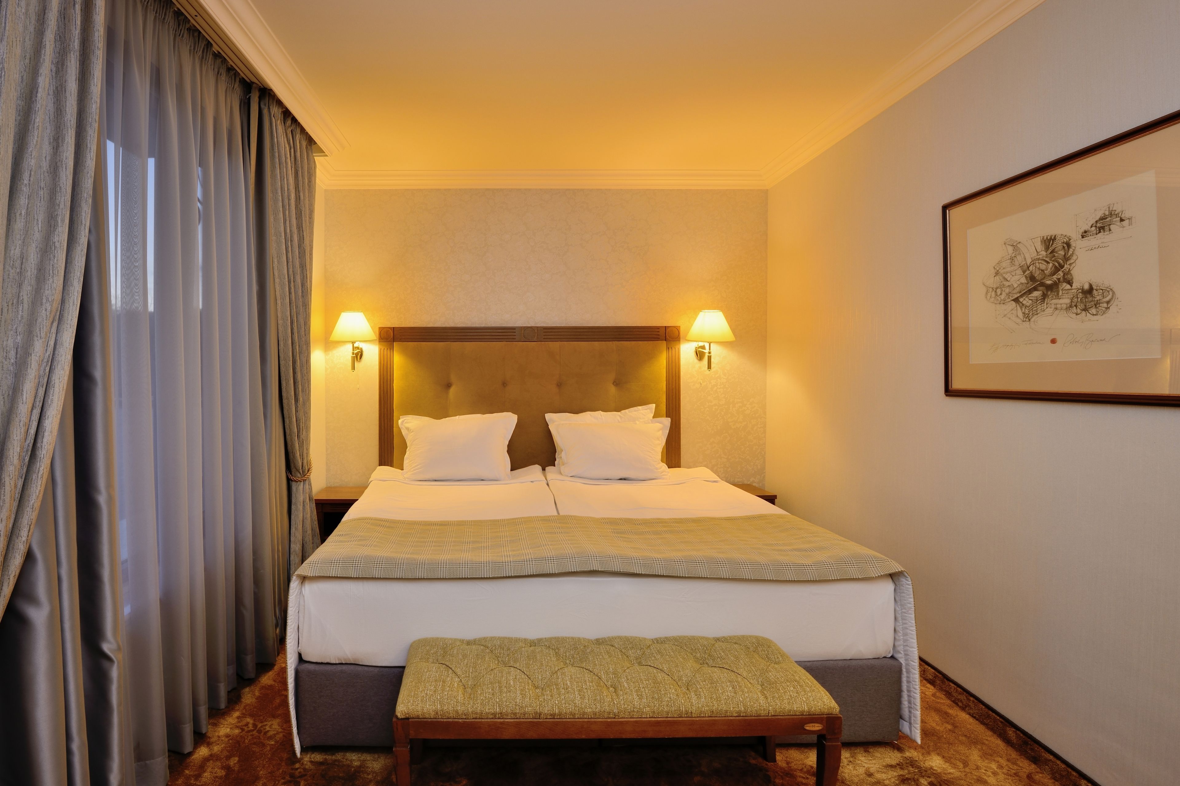 A hotel room with a large bed, two bedside lamps, a framed artwork on the wall, and a window with curtains.