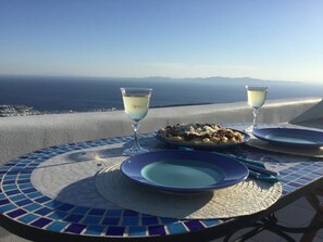 A table with a blue and white mosaic tablecloth, two plates, a bowl of food, and two glasses of white wine, set against a scenic view of the sea and distant mountains. - Blue Azur Deluxe Villa (Tinos)