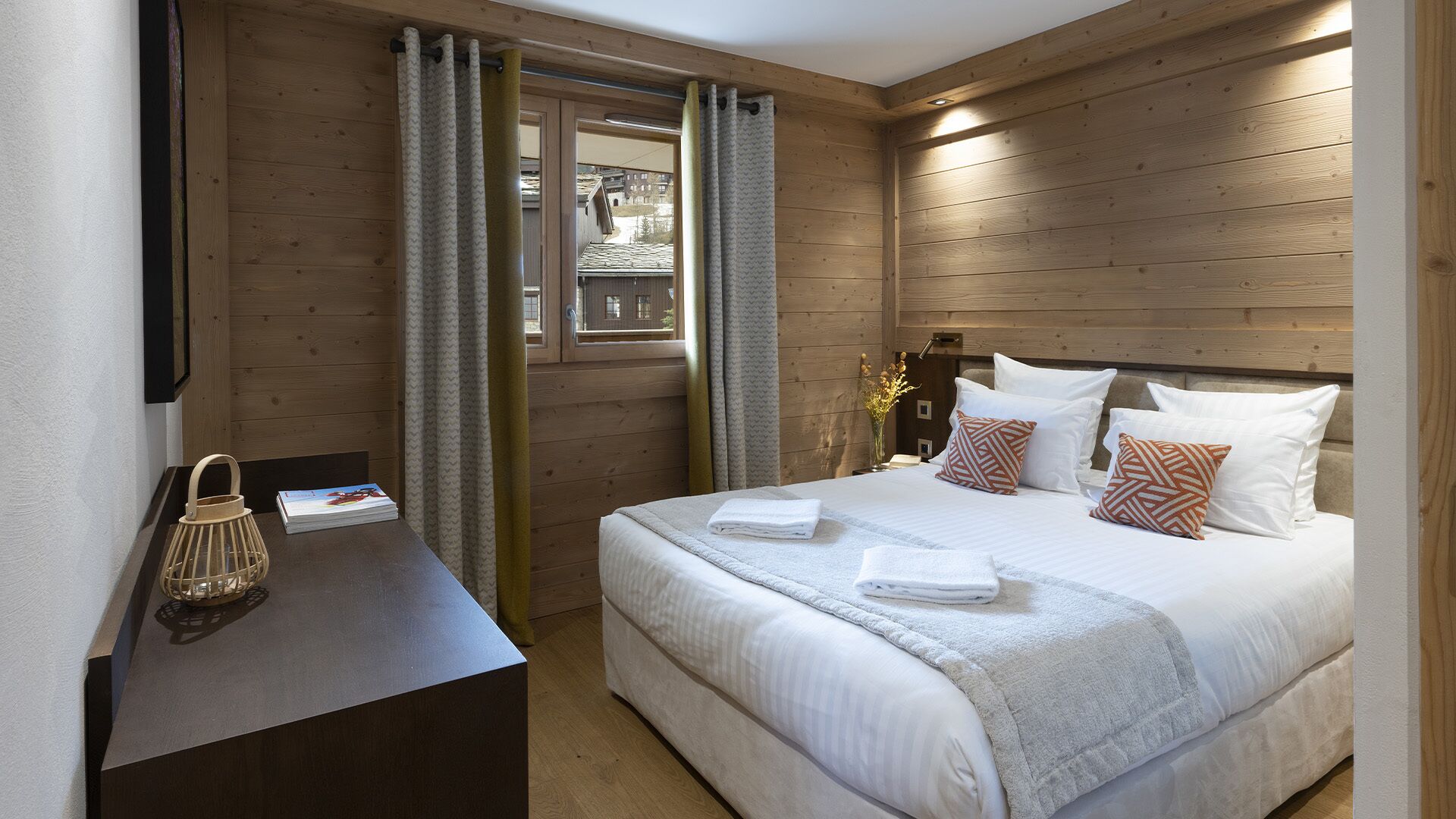 Ein ordentlich bezogenes Bett mit weißen Leinentextilien und gemusterten Kissen, ein Nachttisch mit Lampe und Buch sowie Holzvertäfelung an den Wänden.