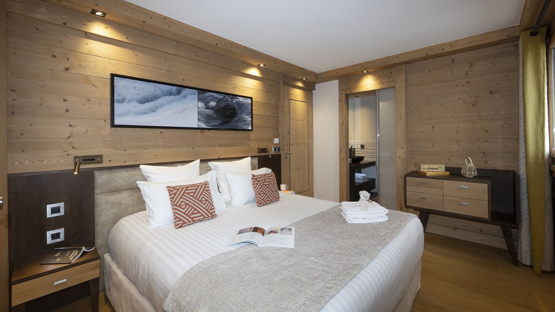 Habitación de hotel con decoración de madera, una cama grande con ropa blanca y almohadas estampadas, una mesita de noche con un libro y un control remoto, otra mesita más pequeña con una taza y un osito de peluche, y un espejo en la pared.