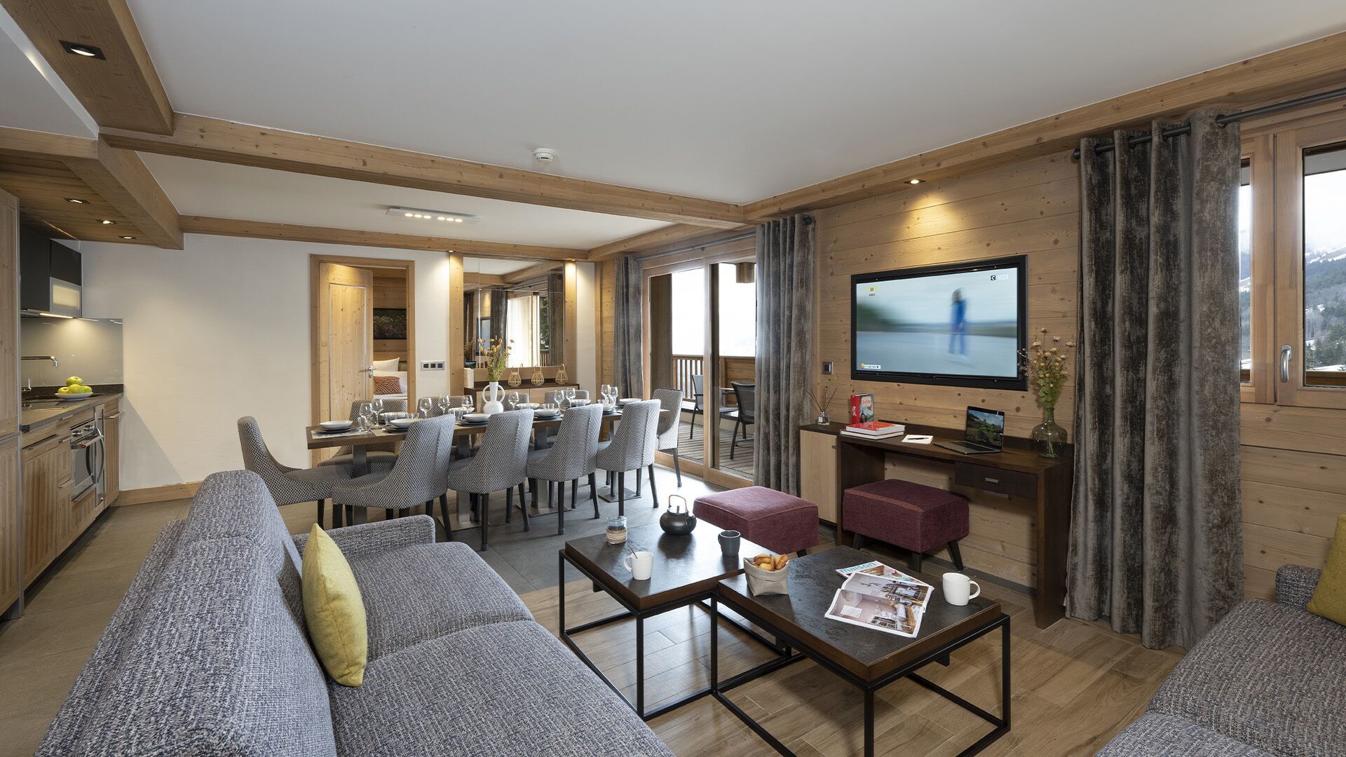 A modern living room with a sofa, coffee table, and TV. There is a dining area with chairs and a table. The room has wooden beams on the ceiling and large windows.