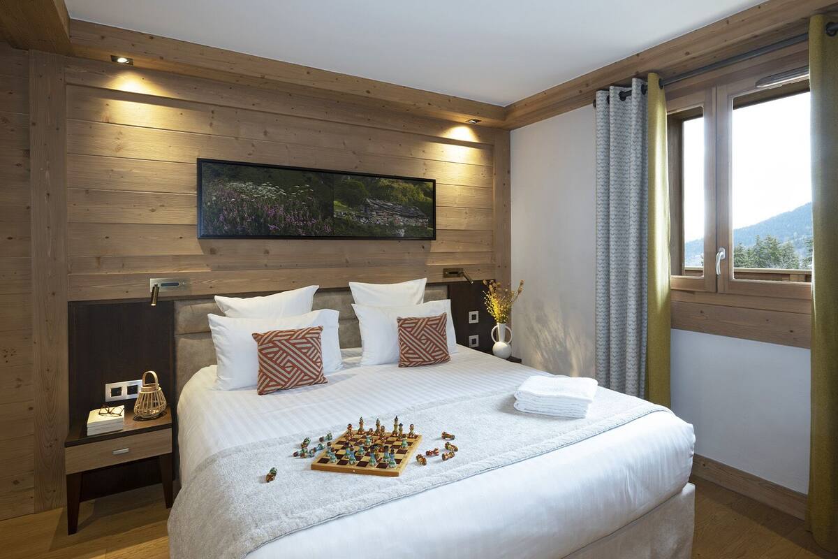 Un dormitorio con techo de madera, una cama grande con ropa blanca, un tablero de ajedrez y una ventana con cortinas.