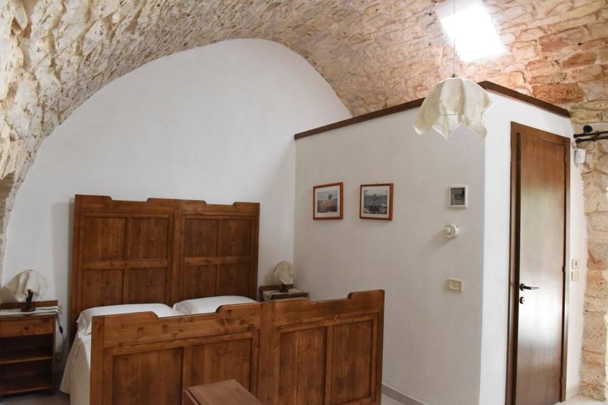 A bedroom with a wooden bed, a nightstand, a desk, and a stone wall.