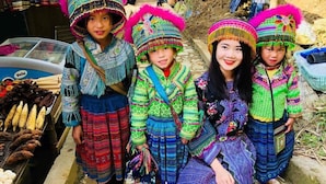 A group of people in traditional attire standing and sitting in a rustic outdoor setting with a market stall in the background. - Sapa Tatu Homestay (Sa Pa)